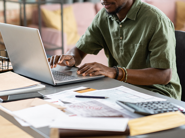 African American man in green shirt sitting at table with papers and examining files on laptop